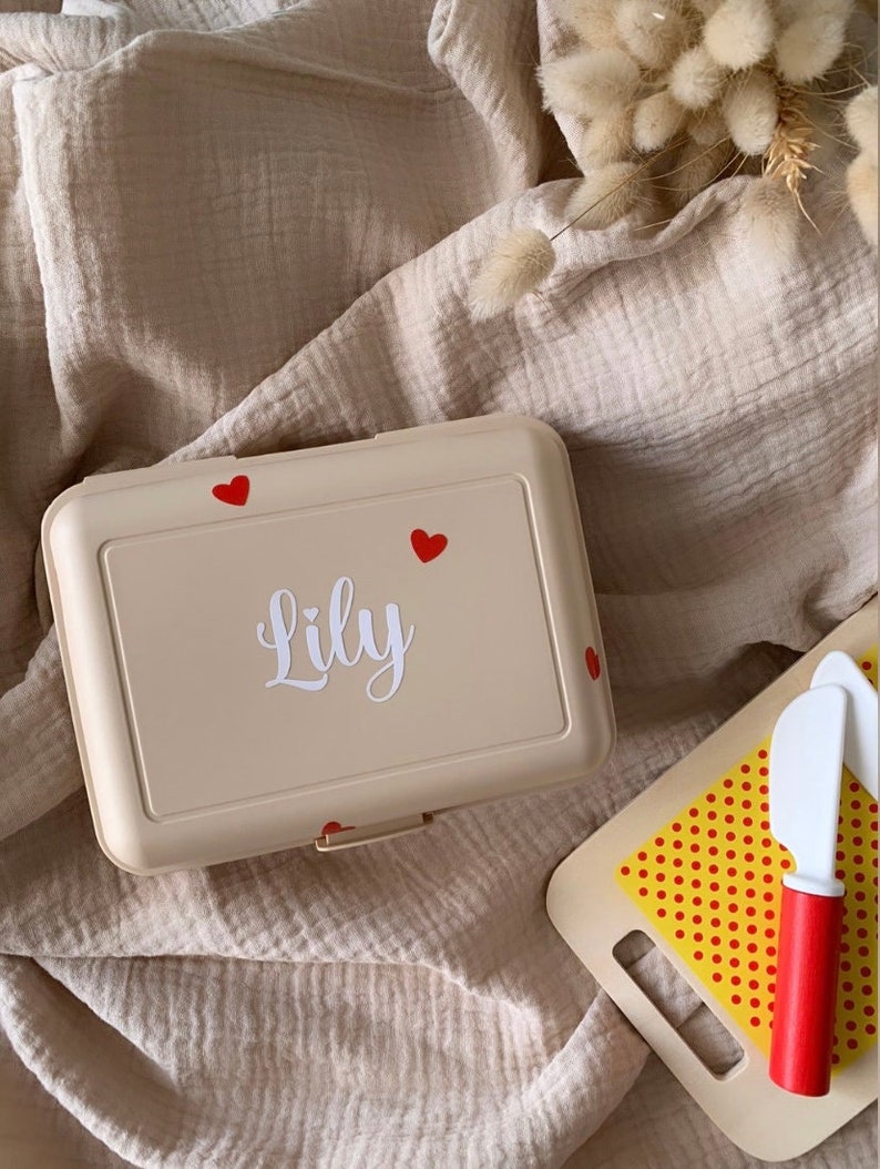 May include: A beige lunchbox with a red heart design and the name "Lily" written in white. The lunchbox is sitting on a beige fabric surface.