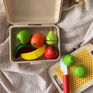 May include: A wooden toy food set with a variety of fruits and vegetables, including a pear, watermelon, banana, strawberry, and lime. The set comes in a beige plastic container with a lid and includes a wooden knife and cutting board with red polka dots.
