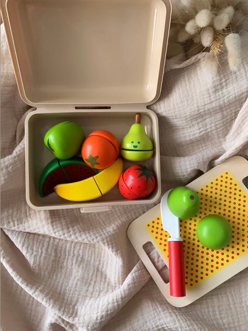 May include: A wooden toy food set with a variety of fruits and vegetables, including a pear, watermelon, banana, strawberry, and lime. The set comes in a beige plastic container with a lid and includes a wooden knife and cutting board with red polka dots.