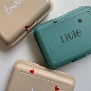 May include: Three beige and teal lunch boxes with red hearts and white names. The names are "&Eacute;milie", "Livio", and "Assiya".