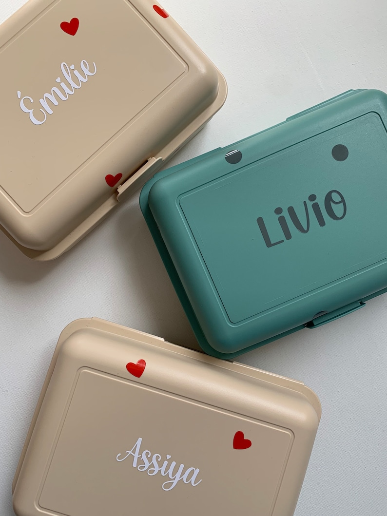 May include: Three beige and teal lunch boxes with red hearts and white names. The names are "&Eacute;milie", "Livio", and "Assiya".