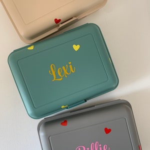 May include: Three lunchboxes with personalized names. The top lunchbox is beige with the name "Tiago" in black lettering and a red heart. The middle lunchbox is green with the name "Lexi" in yellow lettering and yellow hearts. The bottom lunchbox is gray with the name "Billie" in pink lettering and red hearts.