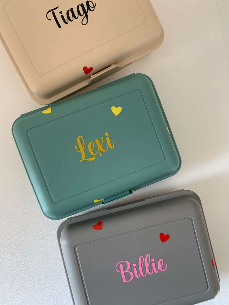 May include: Three lunchboxes with personalized names. The top lunchbox is beige with the name "Tiago" in black lettering and a red heart. The middle lunchbox is green with the name "Lexi" in yellow lettering and yellow hearts. The bottom lunchbox is gray with the name "Billie" in pink lettering and red hearts.