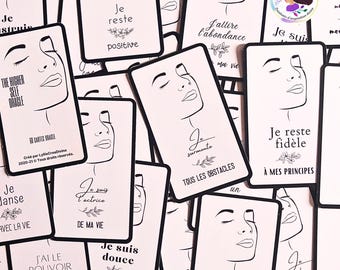 Könnte beinhalten: Eine Sammlung weißer Orakelkarten mit schwarzen Strichzeichnungen von Gesichtern und französischem Text. Jede Karte enthält eine einzigartige Affirmation wie Je reste positive, Je surmonte tous les obstacles und Je suis douce.