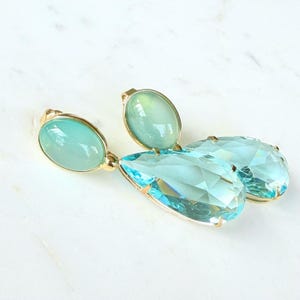 May include: A pair of elegant earrings featuring a gold setting. Each earring has a light blue oval-shaped stone at the top and a large, faceted, teardrop-shaped light blue stone below. The earrings are set against a white background.