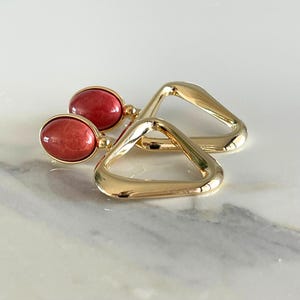 Könnte beinhalten: Ein Paar goldfarbene Ohrringe. Jeder Ohrring hat einen roten, oval geformten Stein und ein dreieckiges geometrisches Design. Die Ohrringe werden auf einer weißen und grauen Marmoroberfläche präsentiert.