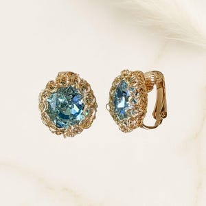 May include: A pair of clip-on earrings featuring large, light blue gemstones. The gemstones are surrounded by a gold-colored, woven metal design. One earring is shown from the side, revealing the clip mechanism.