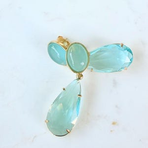 May include: A gold-toned earring featuring three light blue gemstones. The earring has an oval-shaped center stone, a teardrop-shaped stone, and a smaller oval stone. The gemstones are set in gold-colored metal.