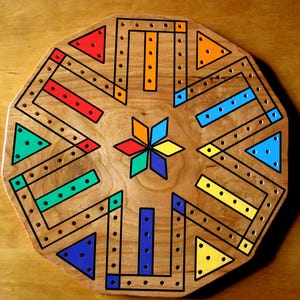 May include: A wooden octagonal game board with a starburst design in the center. The board is painted with black lines and colorful triangles and rectangles. The board has a series of holes around the perimeter.