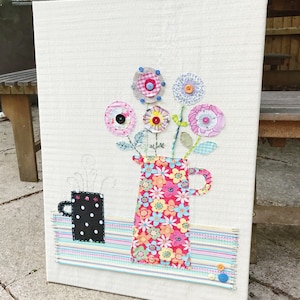 May include: A white canvas featuring a colourful floral arrangement in a red and pink floral patterned jug. The flowers are made of fabric and have buttons in the centre. A black and white polka dot mug with steam coming from it is on a striped surface below the jug.