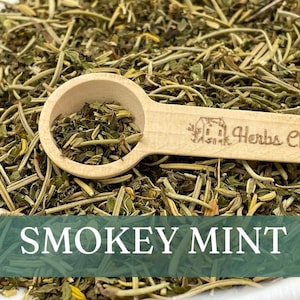 May include: A wooden spoon filled with dried green leafy herbs, labeled "Herbs Chez Moi", sits on a bed of loose dried herbs. The text "SMOKEY MINT" is displayed below the image.