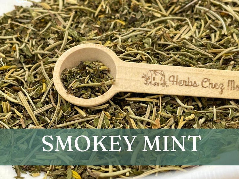 May include: A wooden spoon filled with dried green leafy herbs, labeled "Herbs Chez Moi", sits on a bed of loose dried herbs. The text "SMOKEY MINT" is displayed below the image.