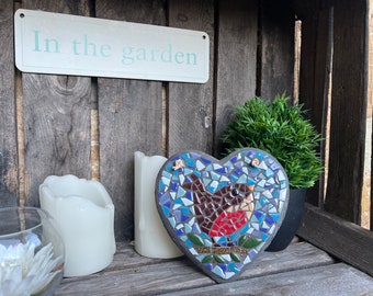 May include: A heart-shaped mosaic with a bird design, featuring blue, brown, and red tiles. The mosaic is displayed with candles, a small potted plant, and a sign that reads "In the garden."