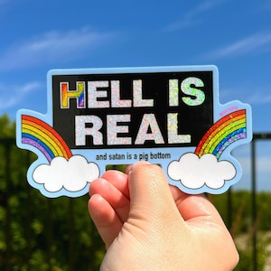 May include: A blue and black sticker with the text "HELL IS REAL" in rainbow glitter letters. The sticker also has two rainbow arcs and clouds with the text "and satan is a pig bottom".