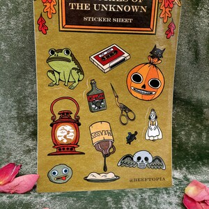 May include: A sticker sheet with a vintage book cover design featuring illustrations of a frog, a cassette tape, a jack-o-lantern, a bottle of liquor, a pair of scissors, a lantern, a bottle of molasses, a turtle, a woman in a dress, a skull with wings, and a rock with a face. The sheet is titled "Memories of the Unknown Sticker Sheet".