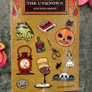 May include: A sticker sheet with a gold background featuring various illustrations, including a frog, a cassette tape, a pumpkin, a lantern, a bottle of molasses, a pair of scissors, a doll, a skull with wings, and a small round creature with a tongue sticking out. The text "Memories of the Unknown" and "Sticker Sheet" are at the top of the sheet. The text "@BEEFTOPIA" is at the bottom of the sheet.