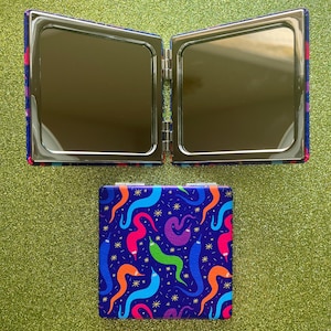 May include: A square, open compact mirror with a vibrant design of colorful worms on a dark blue background. The mirror is open, revealing two reflective surfaces. The compact is on a glittery green surface.