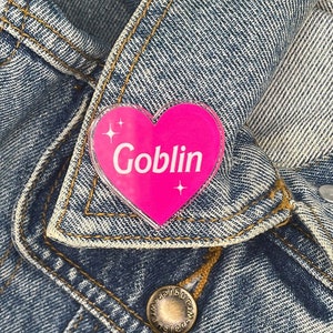 May include: A pink heart-shaped pin with the word "Goblin" in white script and sparkling accents. The pin is attached to a light blue denim jacket. The jacket has visible stitching and metal button details.