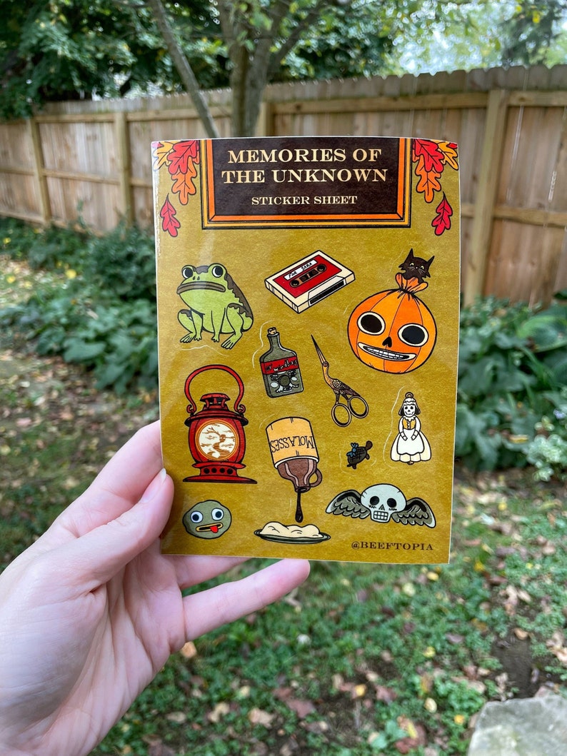 May include: A sticker sheet with a vintage book cover design featuring the text "Memories of the Unknown Sticker Sheet". The sheet includes various stickers with a spooky and whimsical theme, such as a frog, a pumpkin, a lantern, a bottle of molasses, a skull with wings, and a cassette tape.