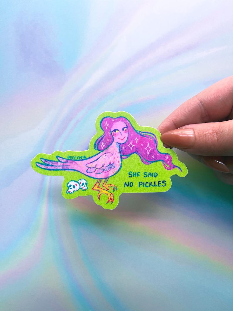 May include: A sticker with a cartoon illustration of a bird-like figure with pink hair and a smiling face. The sticker has a green border and the text "SHE SAID NO PICKLES".
