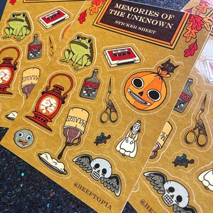 May include: A sticker sheet with a vintage aesthetic featuring illustrations of a skull with wings, a pumpkin with a face, a frog, a bottle of molasses, a lantern, a vintage cassette tape, a pair of scissors, a turtle, a doll, and a bird. The sticker sheet is on a brown background with the text "Memories of the Unknown" and "Sticker Sheet" printed on it.