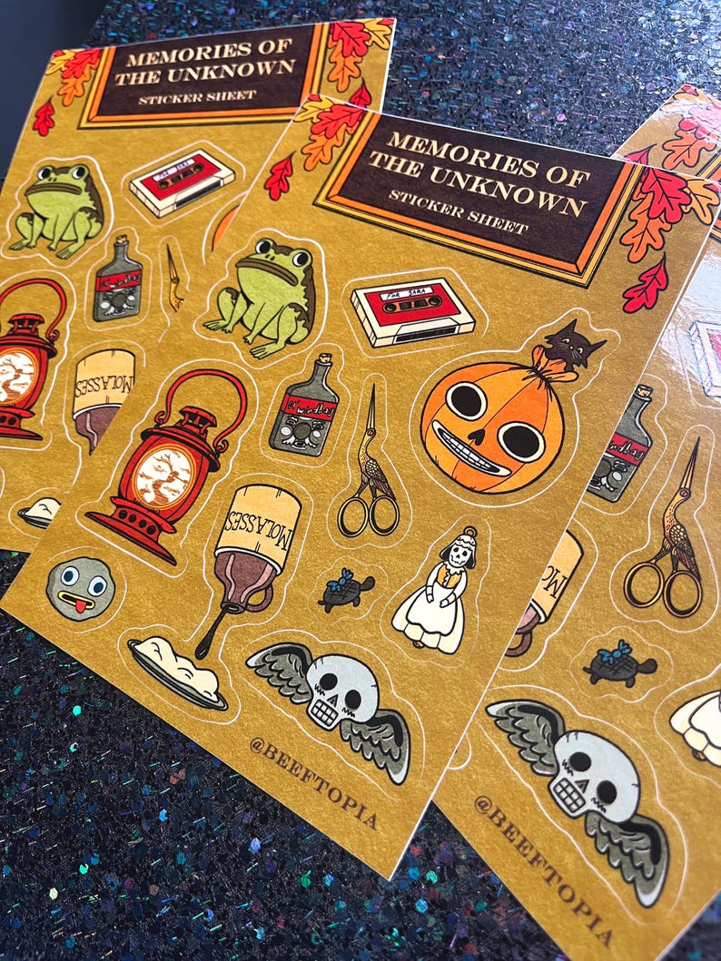 May include: A sticker sheet with a vintage aesthetic featuring illustrations of a skull with wings, a pumpkin with a face, a frog, a bottle of molasses, a lantern, a vintage cassette tape, a pair of scissors, a turtle, a doll, and a bird. The sticker sheet is on a brown background with the text "Memories of the Unknown" and "Sticker Sheet" printed on it.
