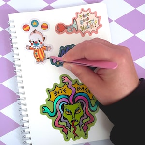 May include: A notebook with colorful stickers featuring a cartoon clown, a snake with the text "Bite Back", and a horn with the text "Toot Your Own Horn!"