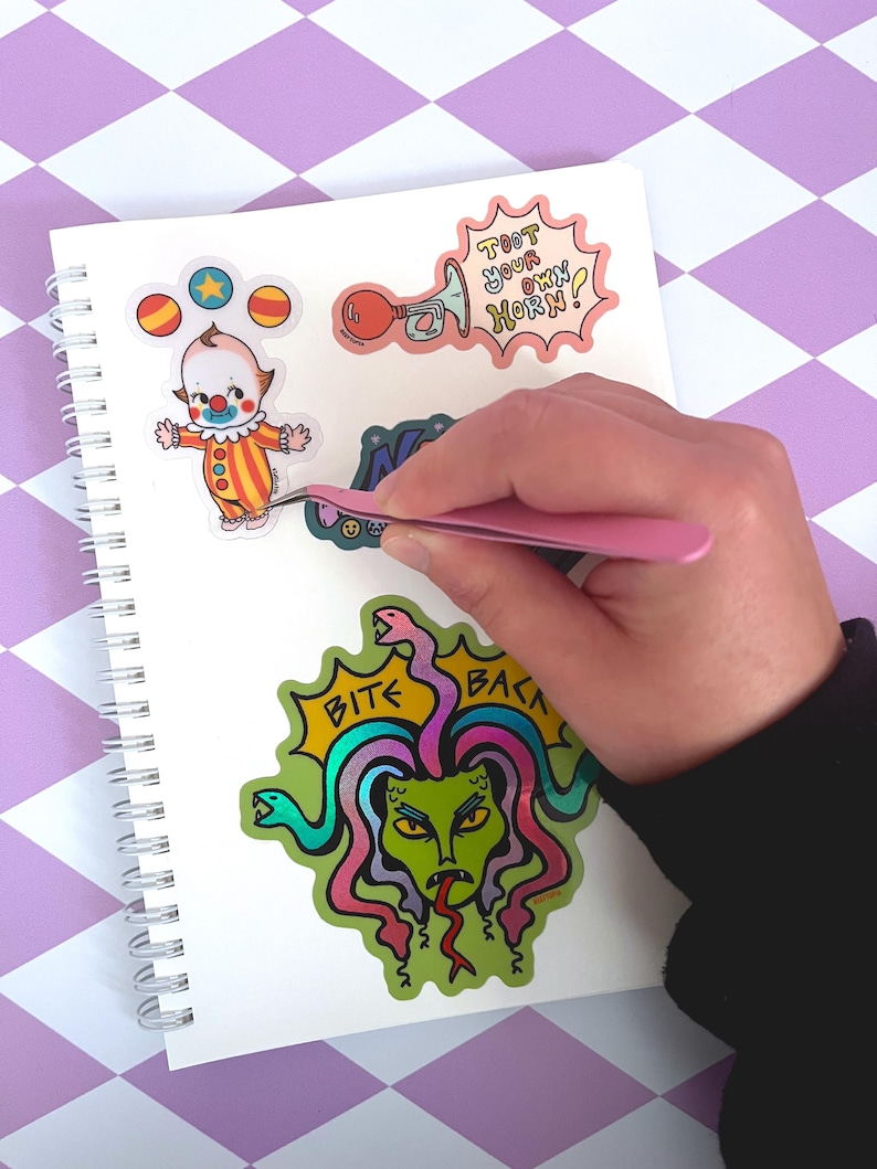 May include: A notebook with colorful stickers featuring a cartoon clown, a snake with the text "Bite Back", and a horn with the text "Toot Your Own Horn!"