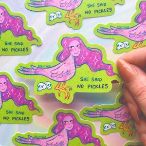 May include: A selection of stickers depicting a pink bird-woman with long, purple hair. Each sticker has a green border and the text "SHE SAID NO PICKLES". The design also includes small skulls. The stickers are displayed on a light background.