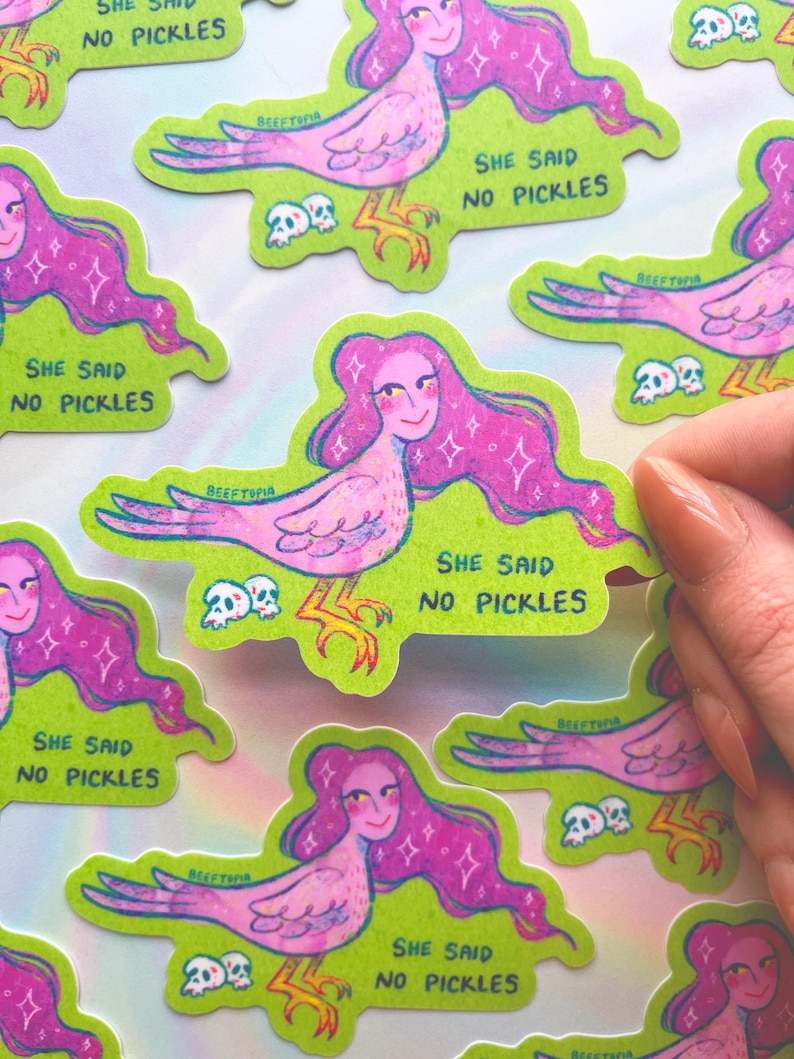 May include: A selection of stickers depicting a pink bird-woman with long, purple hair. Each sticker has a green border and the text "SHE SAID NO PICKLES". The design also includes small skulls. The stickers are displayed on a light background.