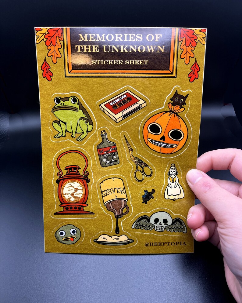 May include: A sticker sheet with a gold background featuring various illustrations, including a frog, a cassette tape, a jack-o-lantern, a bottle of molasses, a lantern, a pair of scissors, a doll, a skull with wings, and a googly-eyed rock. The sheet is titled "Memories of the Unknown Sticker Sheet" and includes the social media handle "@BEEFTOPIA".