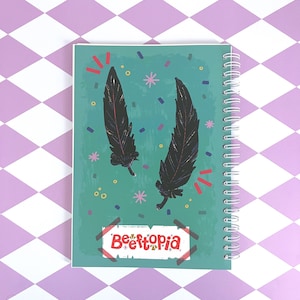 May include: A teal blue notebook with a spiral binding. The cover features two black feathers with glitter accents and confetti in various colors. The text "Befftopia" is written in red and white on a gold sticker.