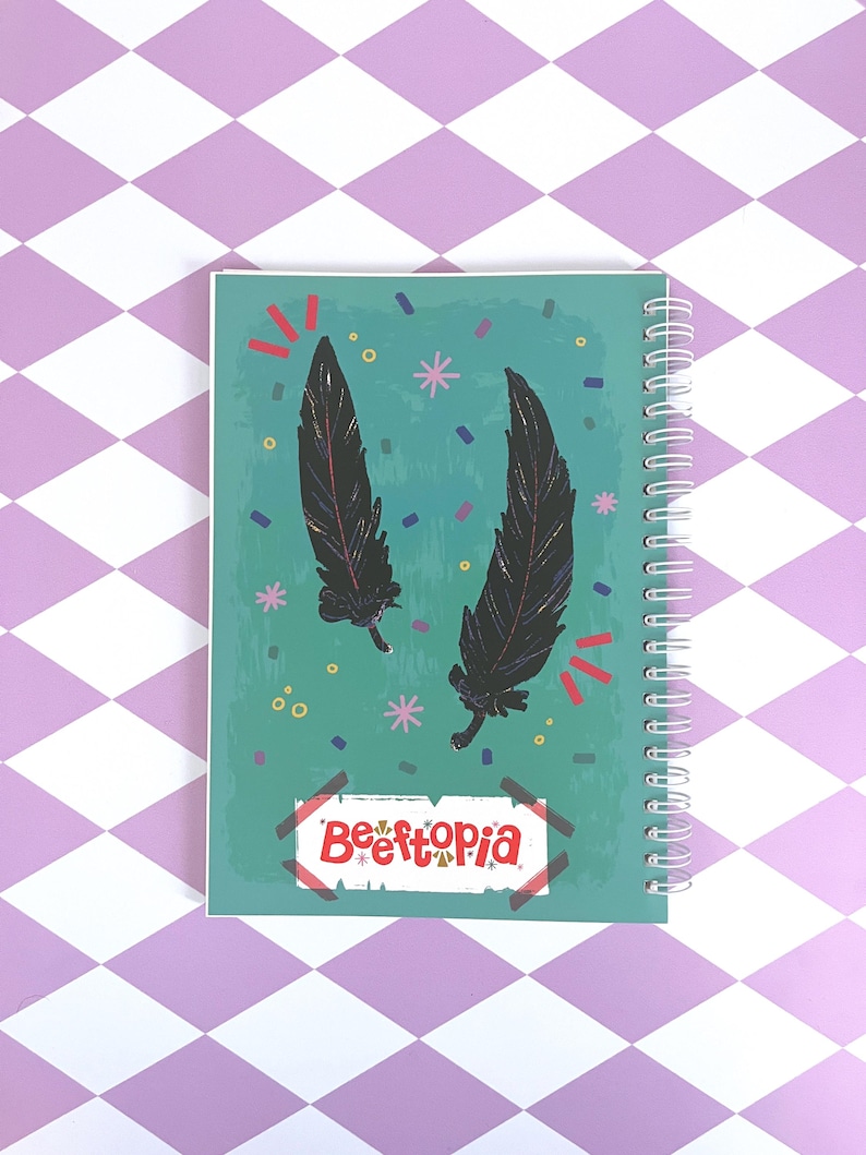 May include: A teal blue notebook with a spiral binding. The cover features two black feathers with glitter accents and confetti in various colors. The text "Befftopia" is written in red and white on a gold sticker.