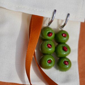 May include: A pair of dangle earrings featuring three stacked green olive beads with red pimento centers on each, suspended from silver-toned metal hoops. The earrings are attached to a cream-colored fabric pouch with the text poom. printed in orange.