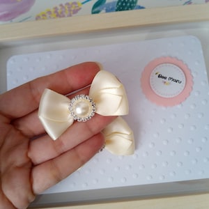 May include: Two ivory satin bows with a pearl and rhinestone embellishment. The bows are displayed on a white background with a pink circle that says "Bee Maru".