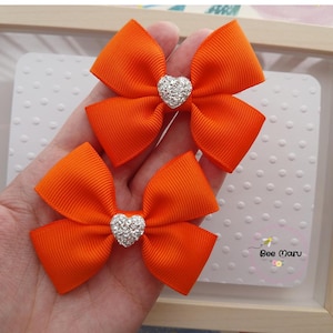 May include: Two bright orange grosgrain ribbon bows, each featuring a central silver-toned heart-shaped embellishment covered in small clear crystals. The bows are constructed with a ribbed texture and are positioned one above the other.