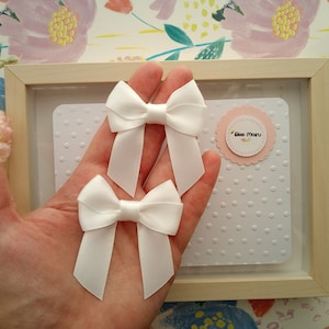 May include: Two white satin bows with a pointed end. The bows are held in a hand against a white background with small polka dots.