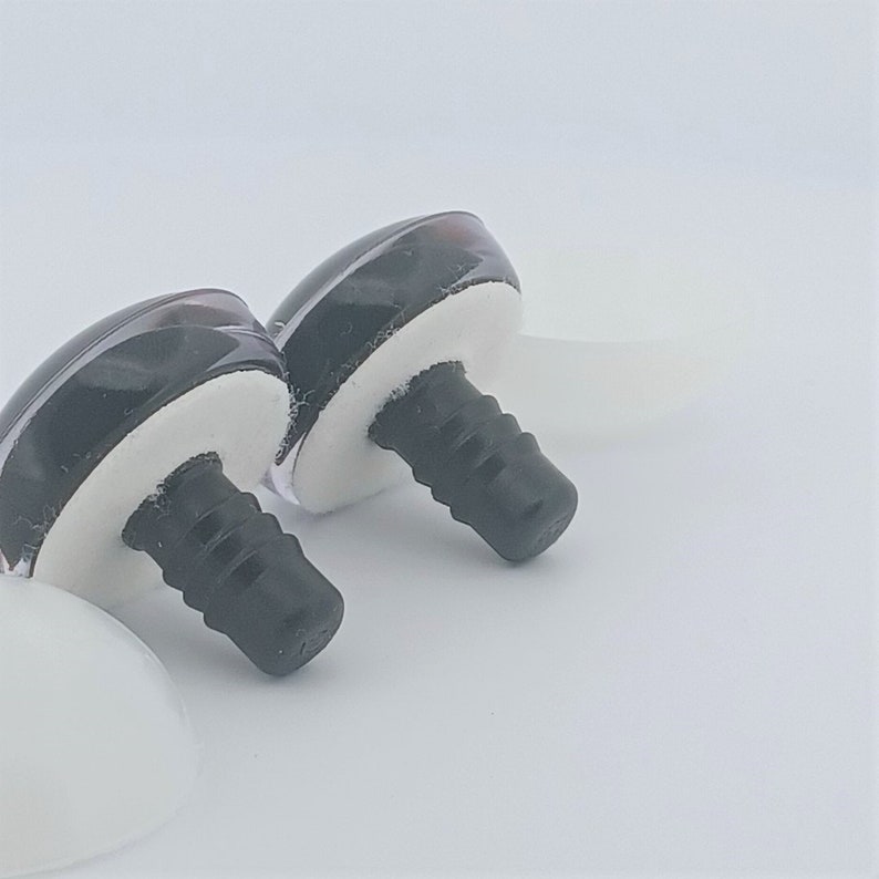 May include: Two black and clear plastic doll eyes with white foam bases and black screw posts. The eyes are glossy and have a reflective quality. The background is plain white.