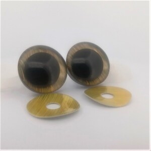 May include: Two large, oval-shaped, brown and black doll eyes with a reflective surface. Two smaller, gold-coloured, tear-drop shaped eye backs with a hole in the centre are also present. The items are set against a white background.
