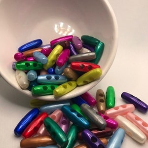 May include: A selection of vibrant, oblong toggle buttons in shades of blue, green, pink, and yellow, spilling from a white ceramic bowl. The buttons are designed with two holes for sewing.