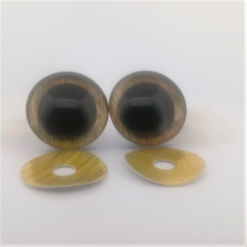 May include: A pair of glossy, black and tan doll eyes with matching tan washers. The eyes have a dark centre and a lighter, swirling pattern around the iris. The washers are oval-shaped with a hole in the centre.