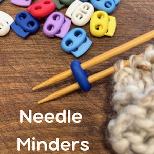 May include: A collection of colorful needle minders in various colors, including blue, purple, red, green, white, and yellow. The image shows the needle minders on a wooden surface, with knitting needles and yarn in the foreground. Text on the image reads "Needle Minders."