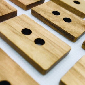 Op de afbeelding: Close-up van verschillende rechthoekige houten knopen met elk twee gaten. De knopen zijn lichtbruin met zichtbare houtnerf en afgeronde randen. De knopen zijn gerangschikt op een witte ondergrond.