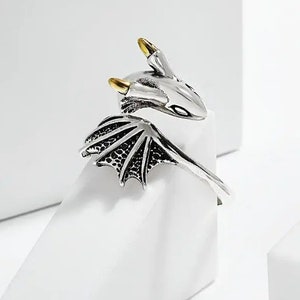 May include: A silver dragon ring with gold-coloured accents on the horns. The ring features a dragon head and wing design. The ring is displayed on a white surface.
