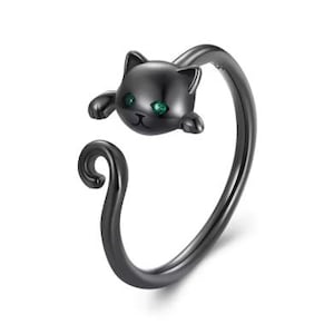 May include: A black cat-shaped ring with green gemstone eyes. The ring features a curled tail design and is made of a dark metal. The cat's head is at the top, with its paws extending down.