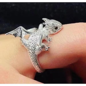 Op de afbeelding: Een zilveren drakenring met gedetailleerde schubben, vleugels en kleine zwarte ogen. De draak wikkelt zich om de vinger en creëert zo een uniek, op fantasie geïnspireerd accessoire. De ring is een sieraad.