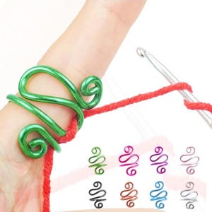 May include: A green metal yarn guide ring worn on a finger, with red yarn threaded through it. Additional rings in green, black, pink, purple, blue, orange, and silver are displayed below. A crochet hook is also visible.