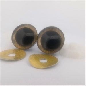 May include: Two large, round, black and tan doll eyes with matching tan eye backing pieces. The eyes have a glossy finish and a detailed, textured appearance. The eye backing pieces are oval-shaped with a hole in the centre.
