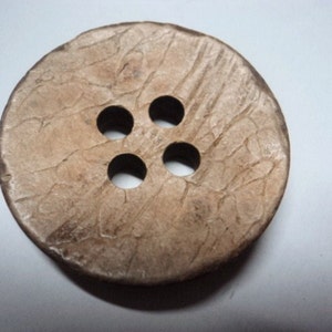May include: A large, round button with four holes. The button is a light brown colour with a textured surface, and appears to be made of a natural material. The button is a simple, classic design.