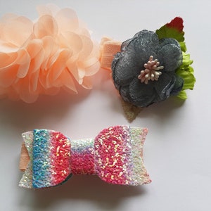 May include: Two elastic headbands and one hair bow. One headband features a large, layered peach-coloured chiffon flower. The second headband has a dark grey fabric flower with green leaves. The hair bow is covered in multi-coloured glitter.
